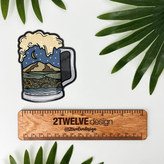 Sticker of a beer mug with Northwest Washington landscape design inside the mug laying on a white background surrounded by leaves and a ruler to determine actual size.