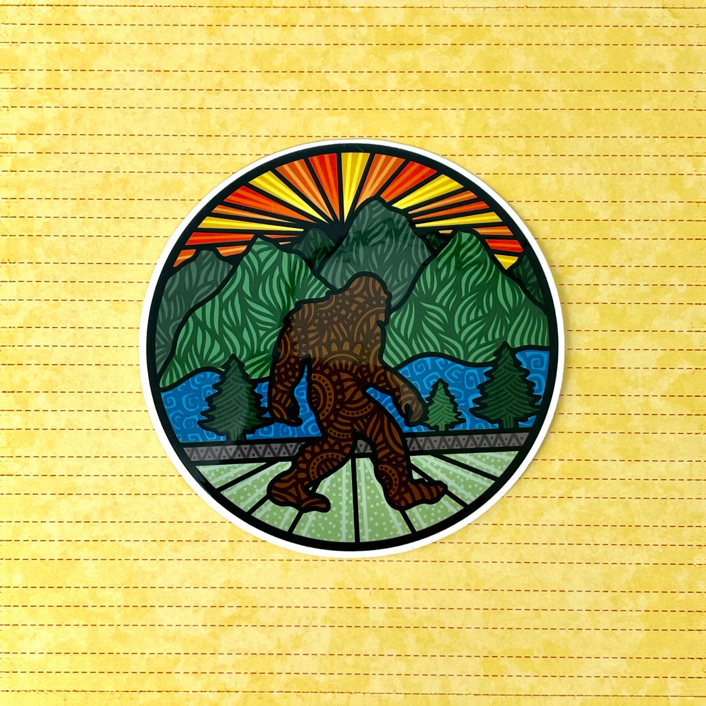 Circular sticker with Bigfoot and a mountain sunrise design laying on a yellow lined background.