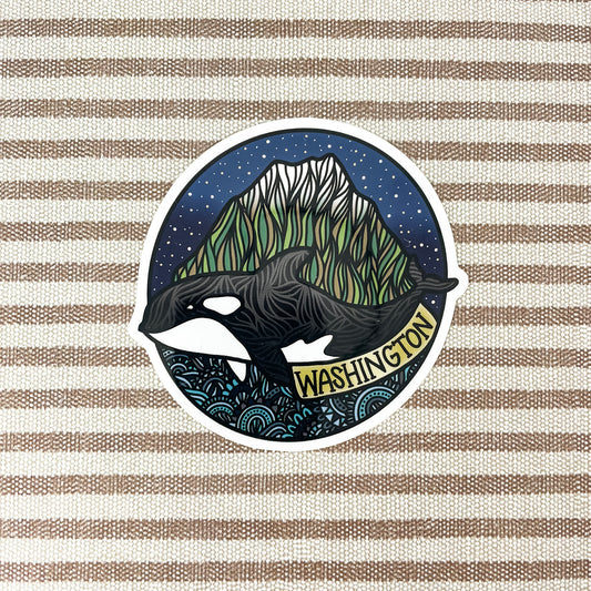 A circular sticker featuring an orca whale in front of a mountain under a starry night sky, with the word 'WASHINGTON' on a banner, shown on a beige and brown striped fabric background.