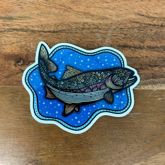 A die-cut sticker featuring an illustrated fish over a blue patterned background, placed on a wooden surface.