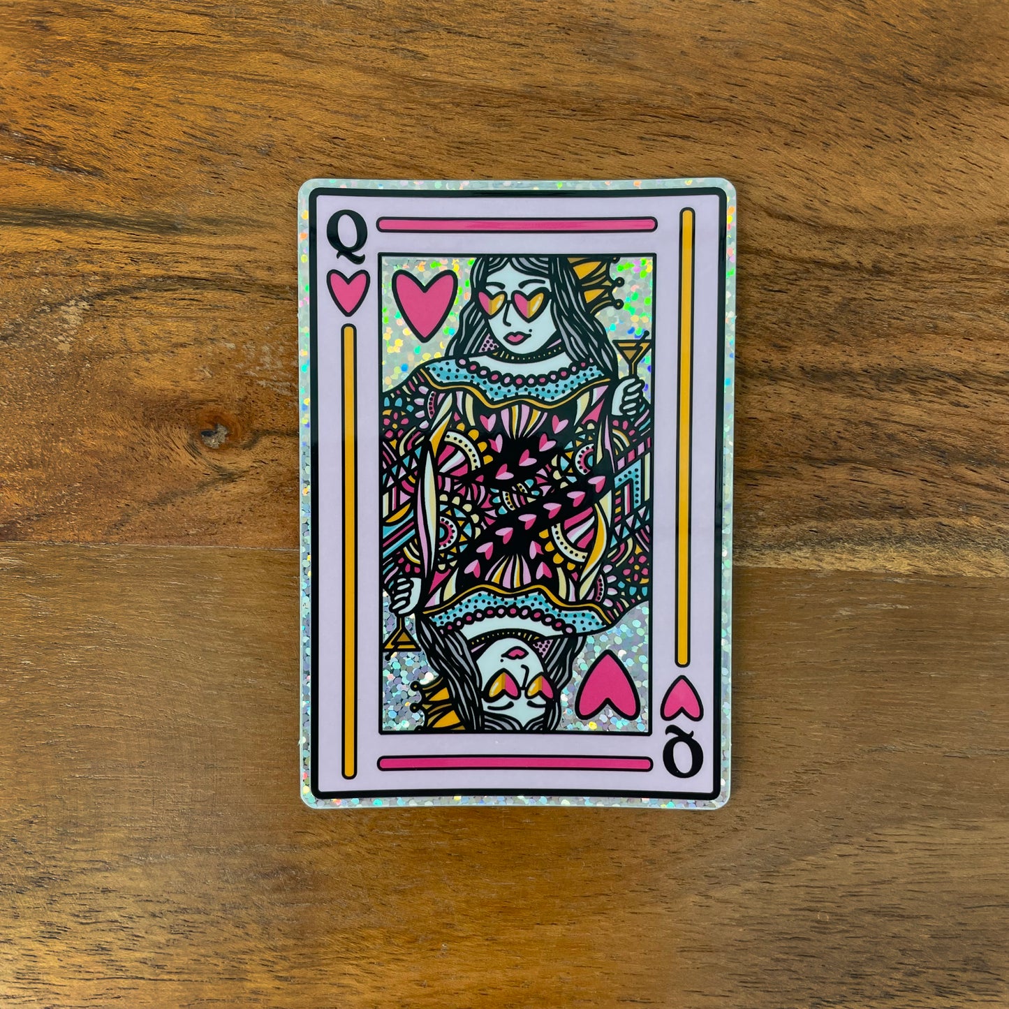 Colorful and glittery Queen of Hearts sticker laying on a wooden surface.
