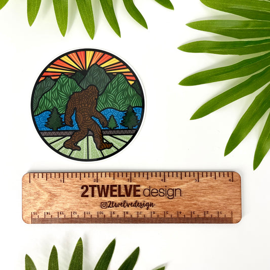 Circular sticker with a stylized mountain and bigfoot illustration. Laying on a white background with leaves surrounding and a ruler to show actual size.