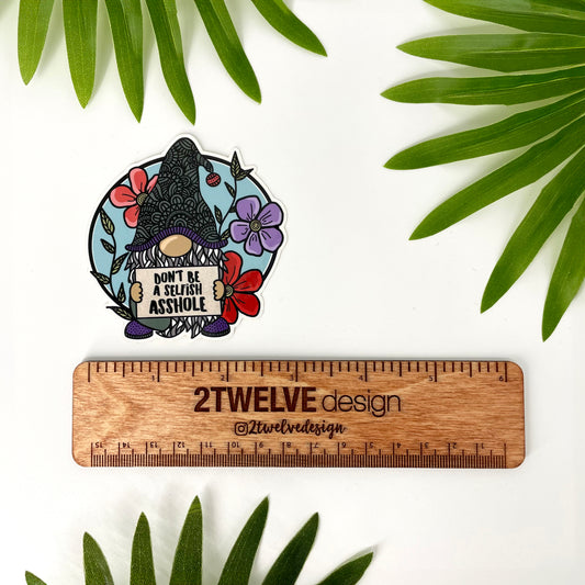 Colorful gnome holding a sign that says "Don't Be A Selfish Asshole". Surrounded by bright flowers with a light blue background. Laying on a white background surrounded by leaves with a ruler to show actual size.