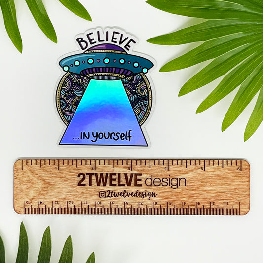 A holographic sticker of a spaceship with the quote "Believe In Yourself" laying on a white background surrounded by leaves with a ruler to show actual size.