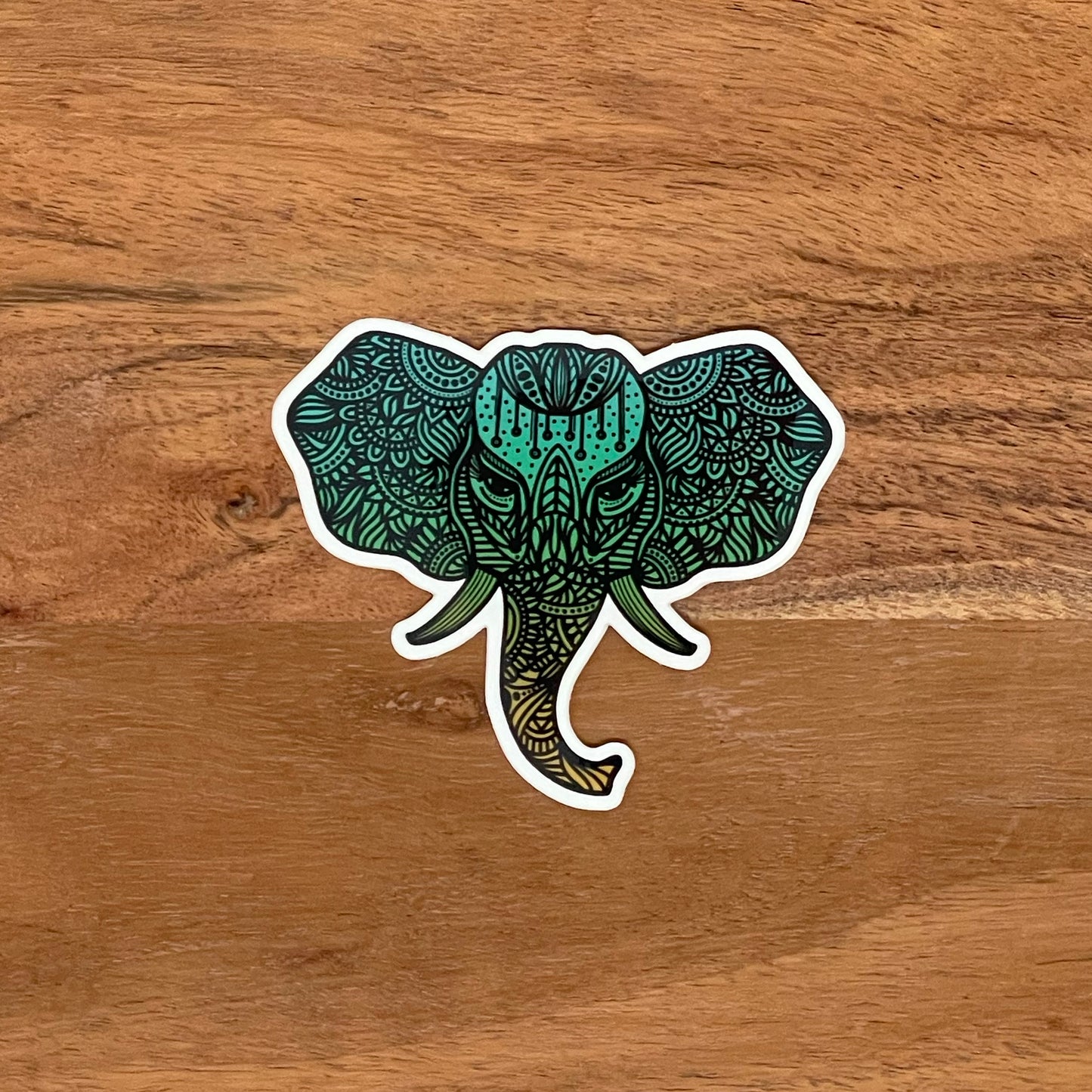 A teal and orange ombre elephant durable sticker. Intricate illustration. Laying on top of a wood surface.
