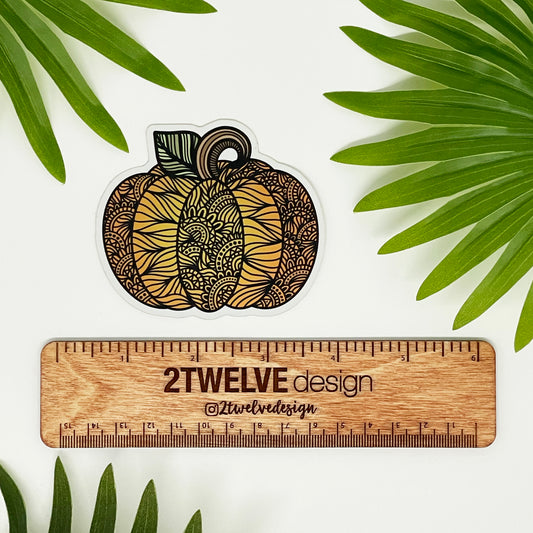 Pumpkin illustration made into a die-cut sticker. Laying on a white background over a wooden "2Twelve Design" ruler. Surrounded by palm leaves.
