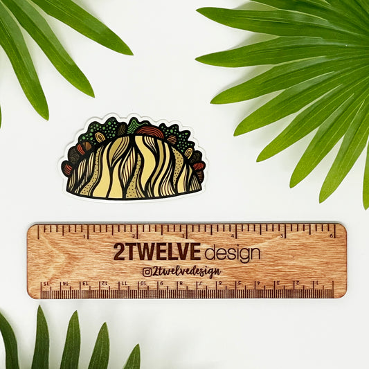 A cute illustrated taco made into a sticker laying on a white surface above a '2Twelve Design' branded ruler, surrounded by palm leaves.
