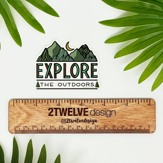 A sticker that reads "Explore The Outdoors" laying on a white background surrounded by leaves and a ruler to show actual size.