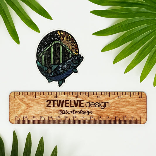 A very detailed fish and mountain illustration made into a die-cut sticker. Laying on a white background above a "2Twelve Design" ruler, surrounded by palm leaves.