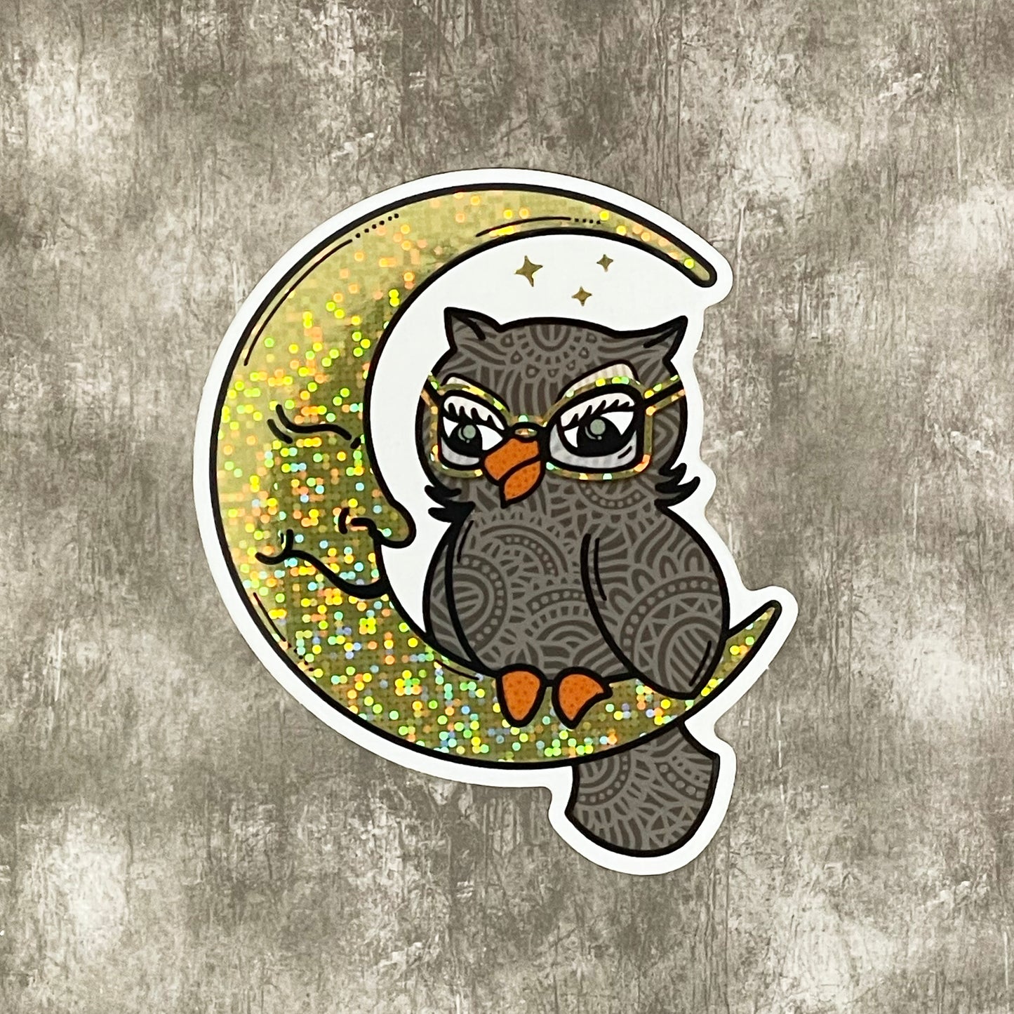 A die-cut glitter sticker featuring a hand-drawn owl and moon illustration with gold glitter accents. Laying on a textured gray background.