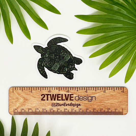 A green turtle sticker laying on a white surface above a ruler that says "2Twelve Design" surrounded by palm leaves