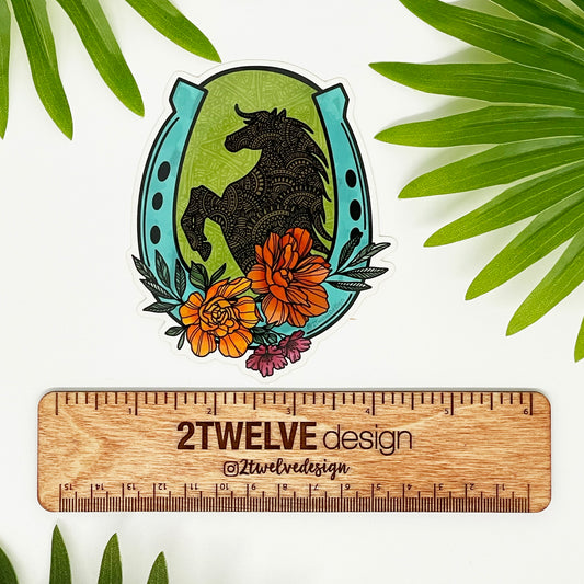 A vinyl, waterproof die-cut sticker featuring a hand-drawn floral and horse illustration, with a horseshoe shape at the top, placed on a wooden surface with green foliage in the background.