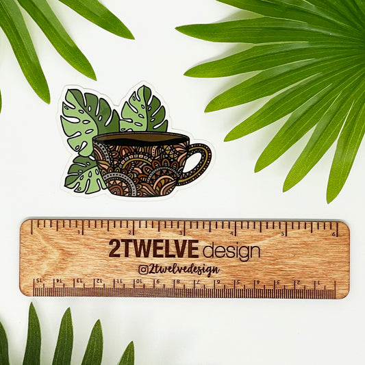 A die-cut vinyl sticker featuring a hand-drawn illustration of a coffee mug with monstera leaves laying on a white background above a "2Twelve Design" ruler, surrounded by palm leaves.