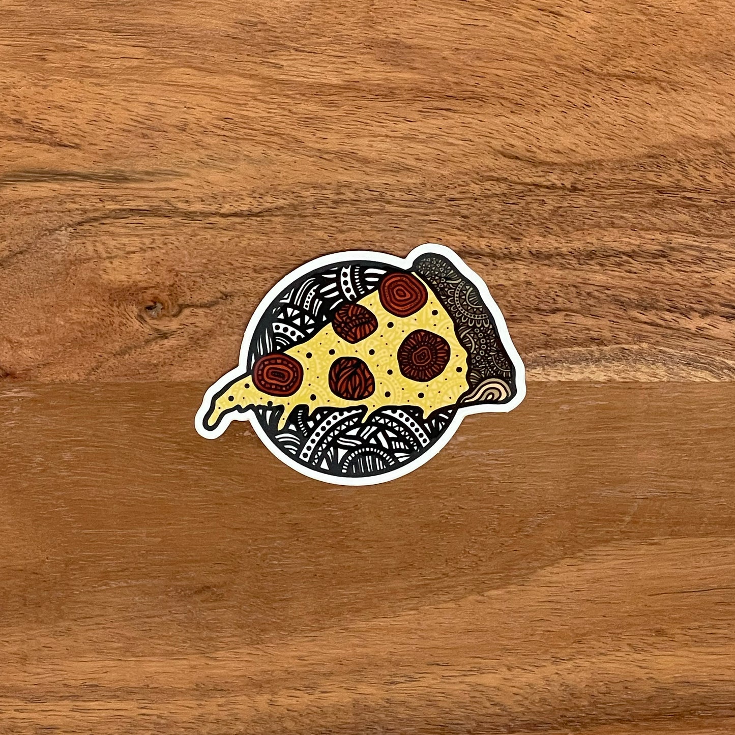 A hand drawn pizza illustration made into a die-cut pizza sticker. Laying on a wooden surface.
