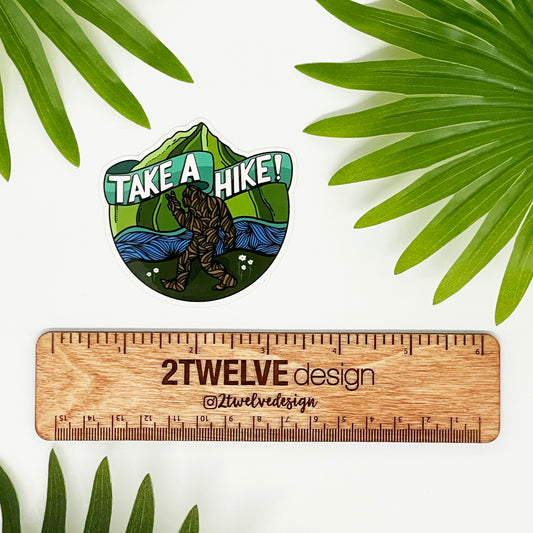 A sticker with bigfoot walking in front of mountains and water. Surrounded by leaves and a ruler to show actual size of sticker.