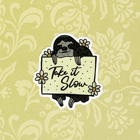 A sticker of a sloth holding a sign with "Take It Slow" written on it. Laying on a light yellow background.