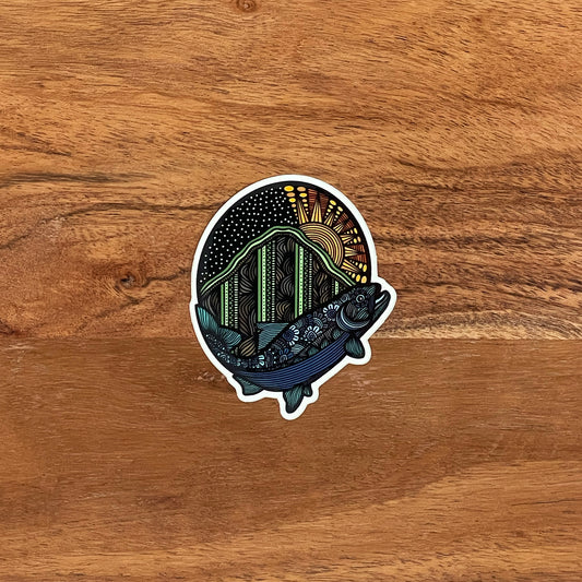A detailed trout and mountain illustration made into a die-cut sticker. Laying on a wooden background.