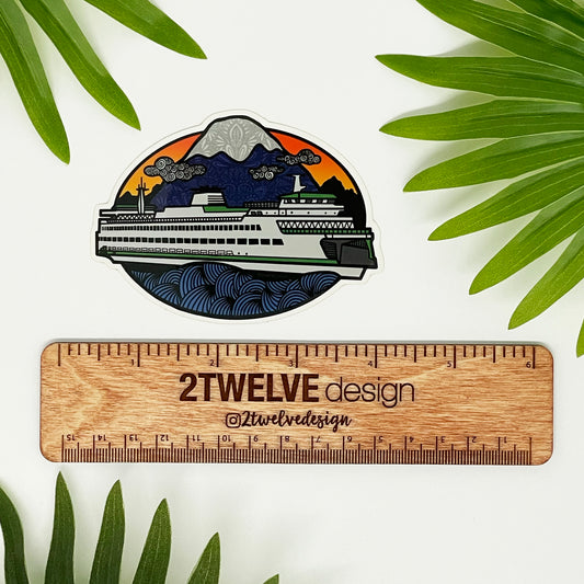 A diecut vinyl sticker of an illustrated ferry boat in Washington State. Laying on a white background above a wood "2Twelve Design" ruler, surrounded by palm leaves.