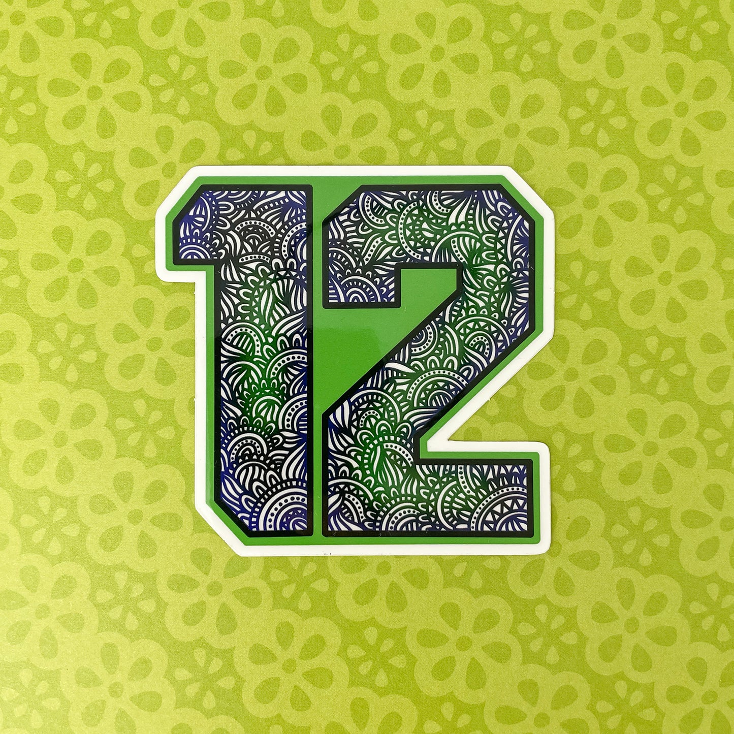 A stylized number 12 with a green, blue and black ornamental patterned fill, outlined in white, centered on a light green patterned background.