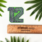 A die-cut sticker featuring the number 12 filled with a green, blue, and white wave pattern, displayed above a wooden ruler engraved with '2Twelve Design'. The scene is styled with green palm leaves on a white background.