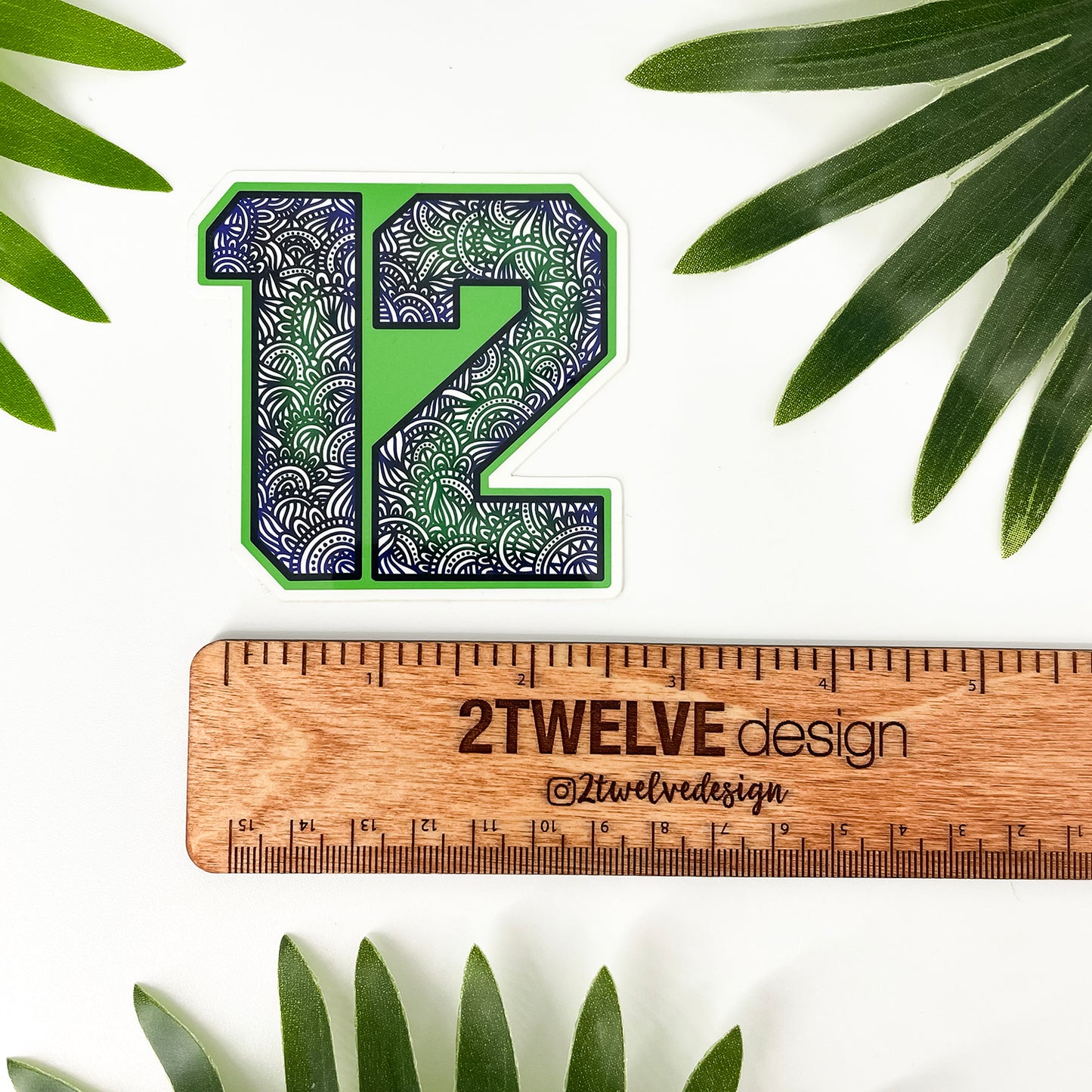 A die-cut sticker featuring the number 12 filled with a green, blue, and white wave pattern, displayed above a wooden ruler engraved with '2Twelve Design'. The scene is styled with green palm leaves on a white background.