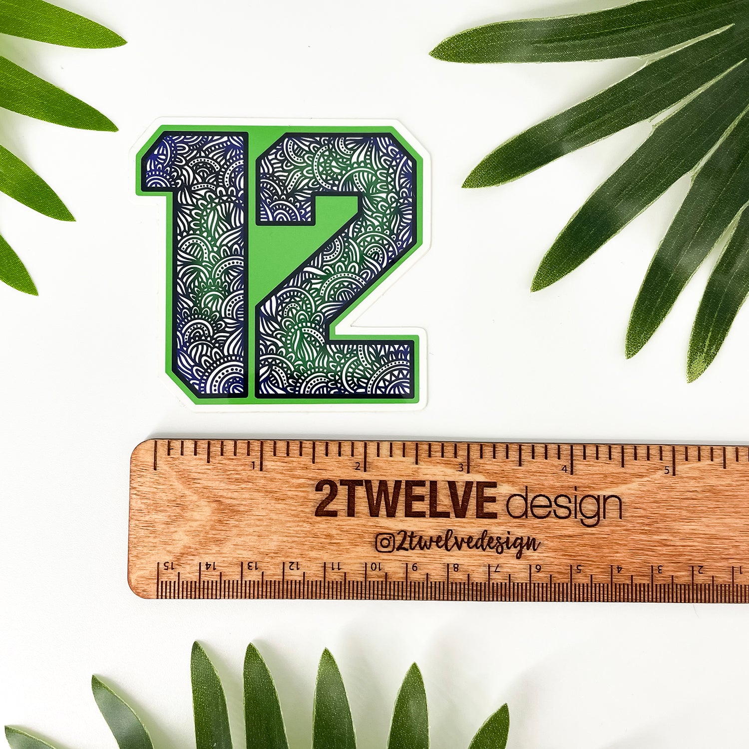 A die-cut sticker featuring the number 12 filled with a green, blue, and white wave pattern, displayed above a wooden ruler engraved with '2Twelve Design'. The scene is styled with green palm leaves on a white background.