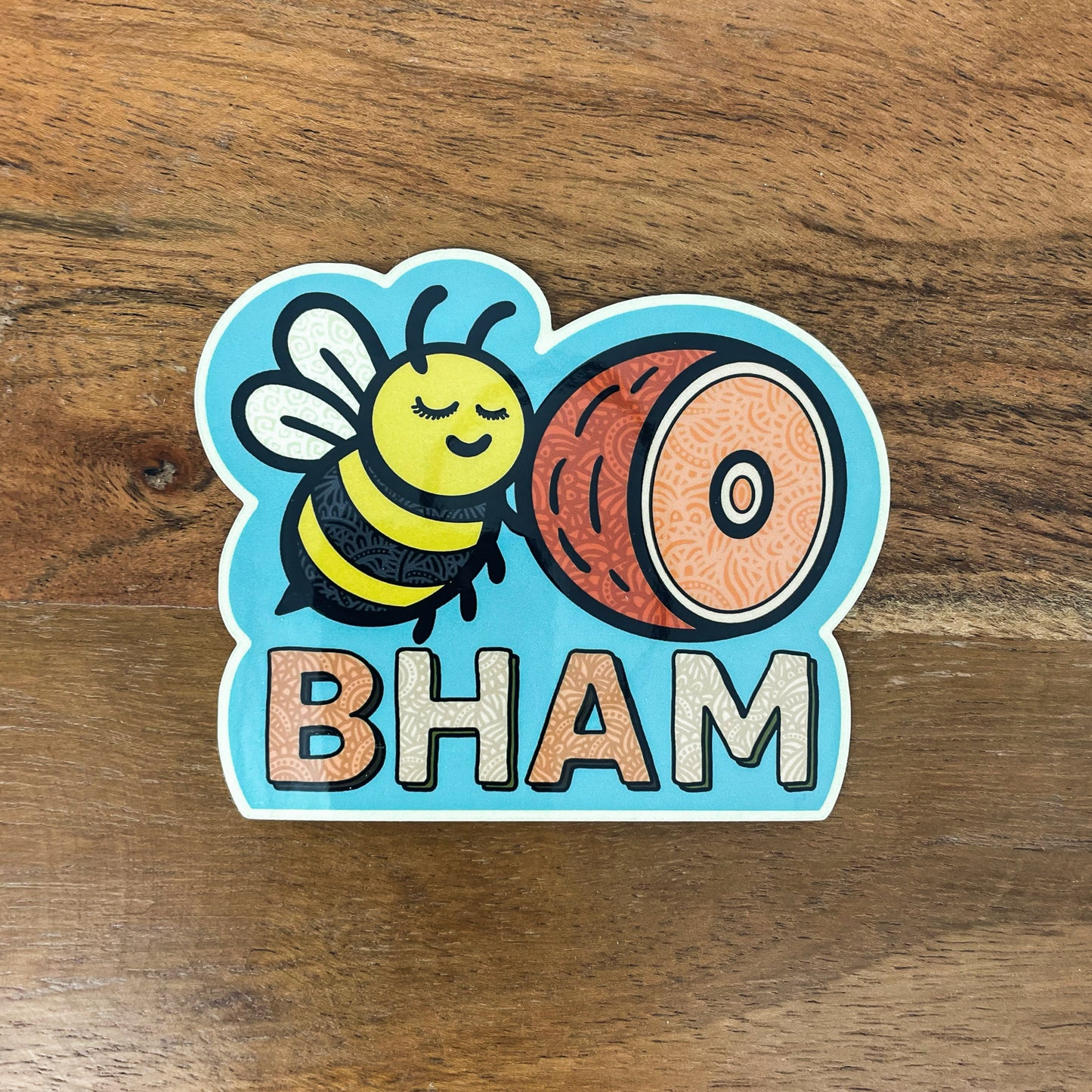 Sticker with a bee and ham illustration labeled 'BHAM' laying on a wooden surface. Bellingham, Washington sticker.