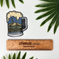 Sticker of a beer mug with Northwest Washington landscape design inside the mug laying on a white background surrounded by leaves and a ruler to determine actual size.