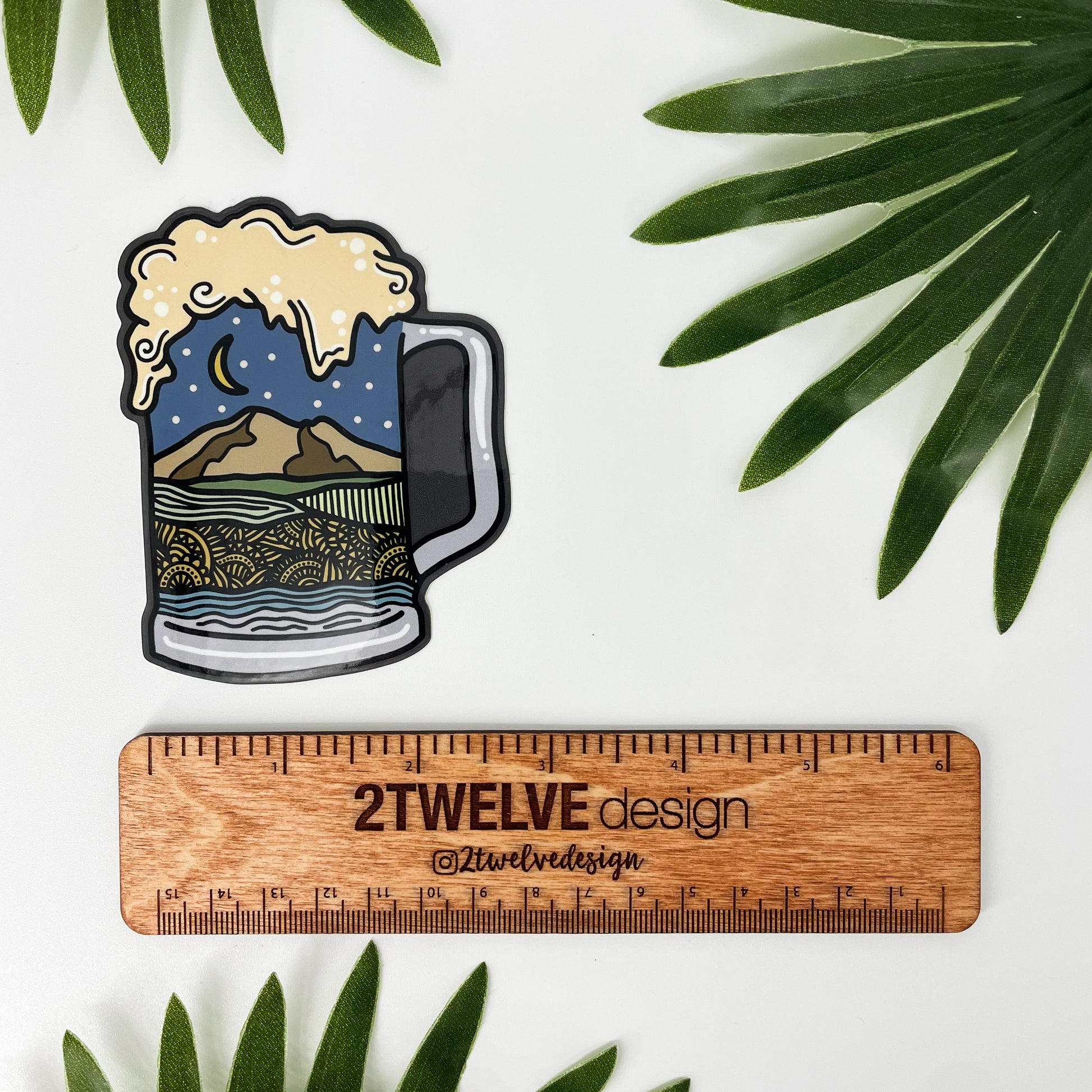 Sticker of a beer mug with Northwest Washington landscape design inside the mug laying on a white background surrounded by leaves and a ruler to determine actual size.