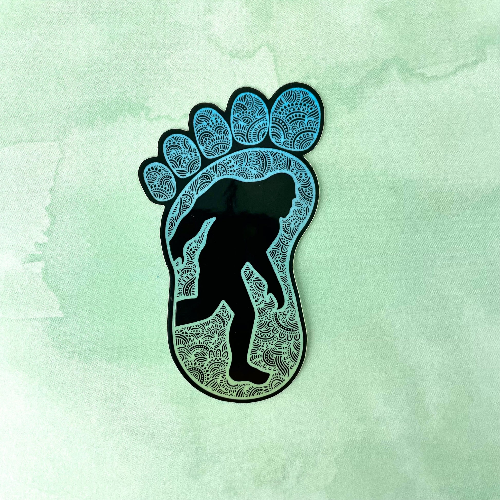 Decorative Bigfoot sticker in a foot shape. Laying on top of a light green texture background.