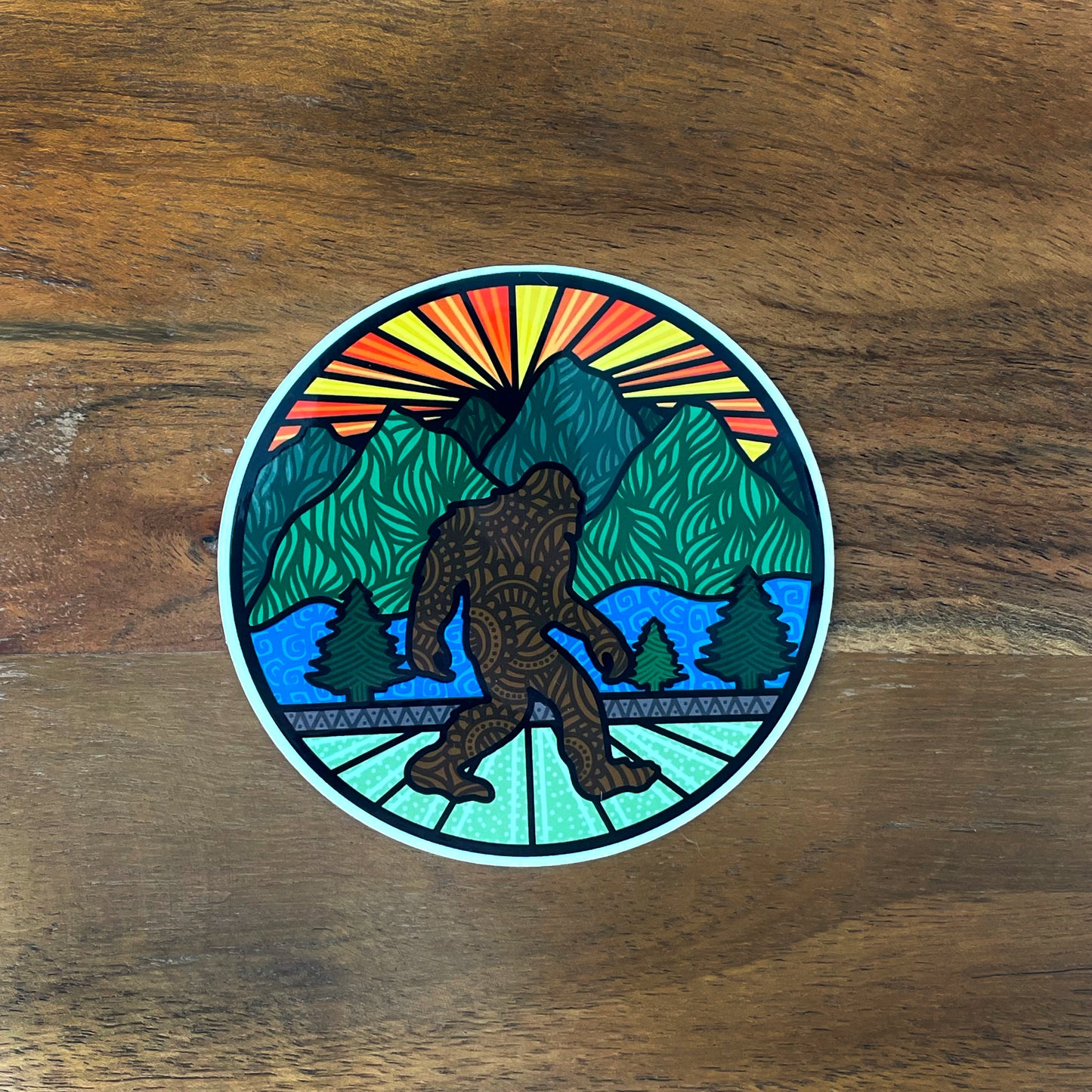 Round sticker with Bigfoot and sunset landscape illustration. Laying on a wood surface.