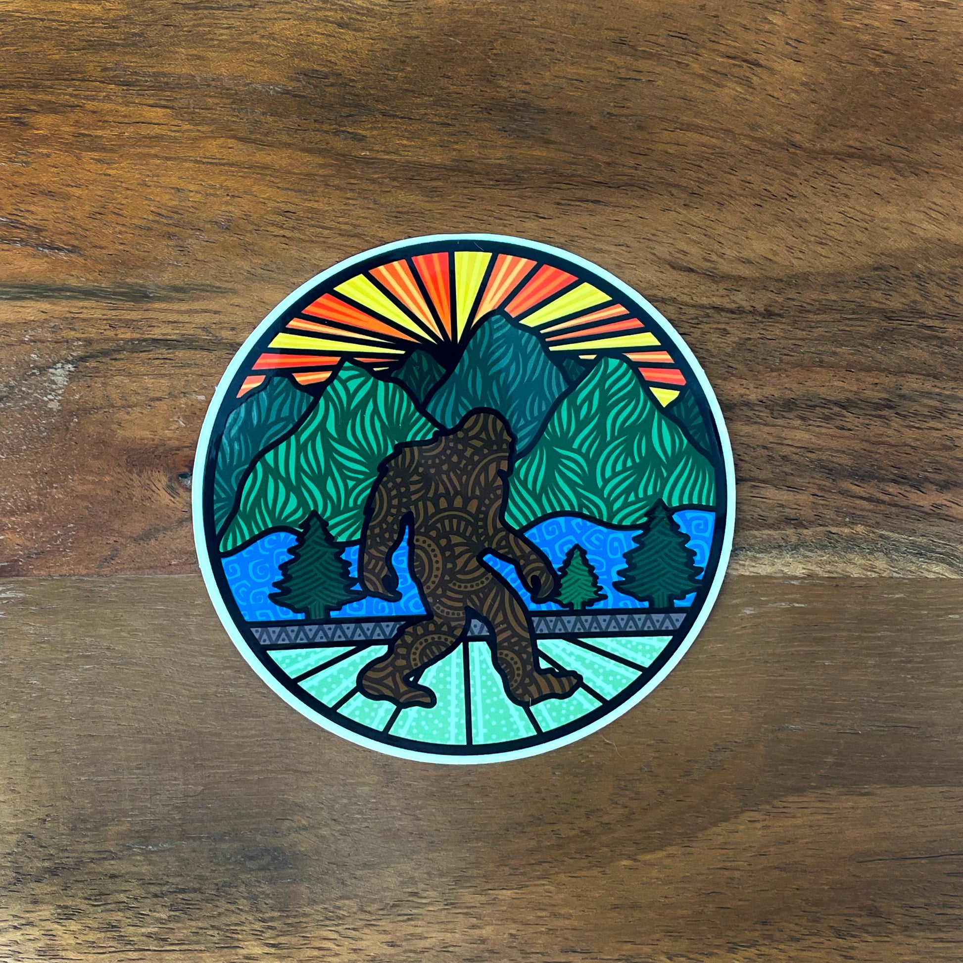 Round sticker with Bigfoot and sunset landscape illustration. Laying on a wood surface.