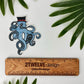Blue kraken/octopus with a top hat and mustache. Detailed illustration made into a vinyl sticker laying on a white background surrounded by leaves and a rule to show actual size.