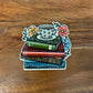 Sticker of a stack of books with a teacup and flowers on a wooden surface.