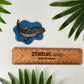 A fish-shaped sticker with a bubbly background and a detailed and colorful trout illustration, shown above a wooden ruler labeled '2Twelve Design'. Green leaves are placed around the edges on a white surface.