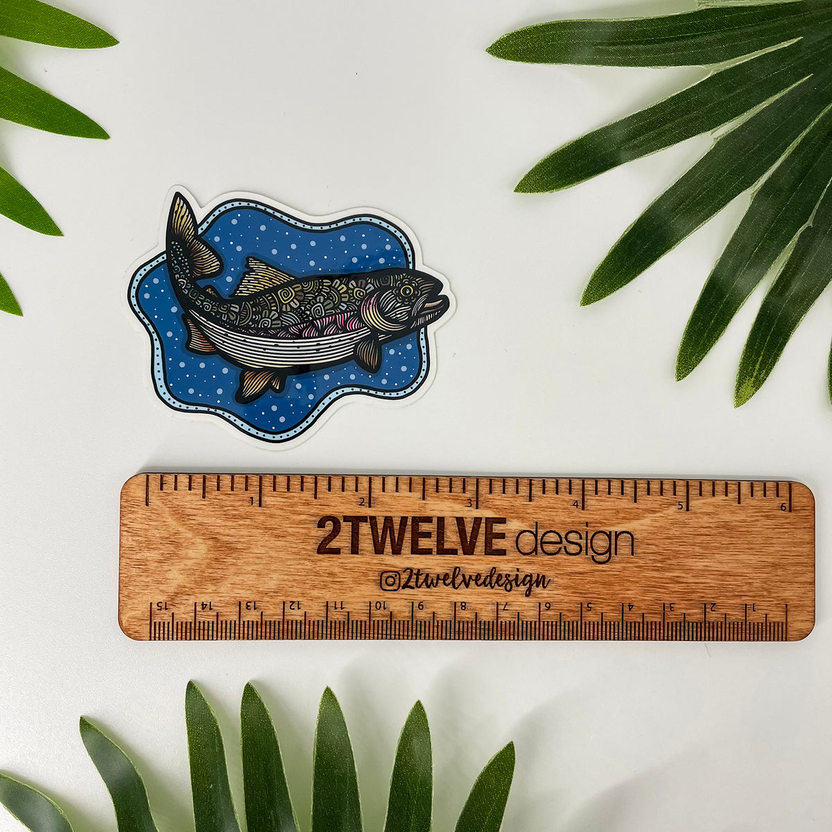 A fish-shaped sticker with a bubbly background and a detailed and colorful trout illustration, shown above a wooden ruler labeled '2Twelve Design'. Green leaves are placed around the edges on a white surface.