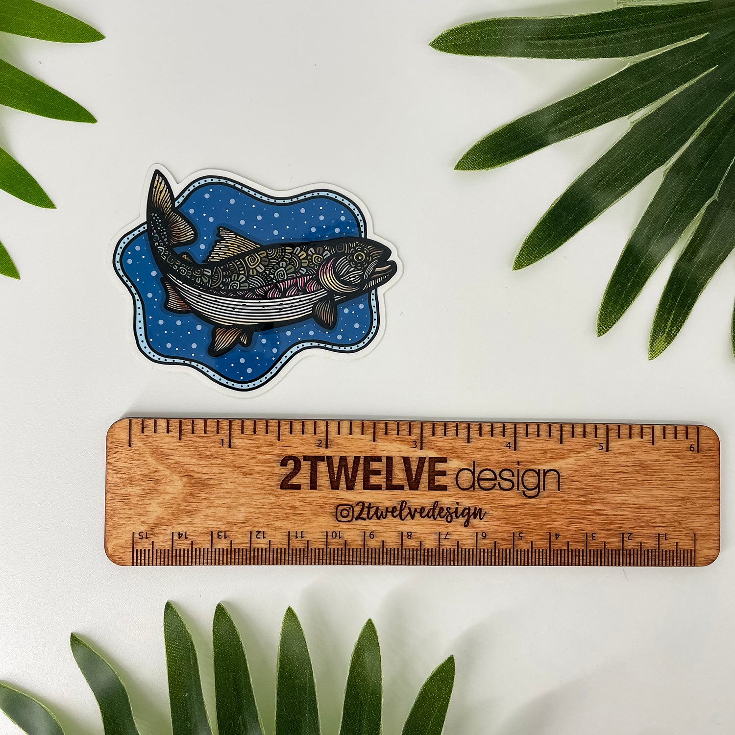 A fish-shaped sticker with a bubbly background and a detailed and colorful trout illustration, shown above a wooden ruler labeled '2Twelve Design'. Green leaves are placed around the edges on a white surface.