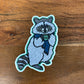 A die-cut sticker of a raccoon wearing a blue striped scarf holding a mug with 'PNW' written on it, placed on a wooden surface.