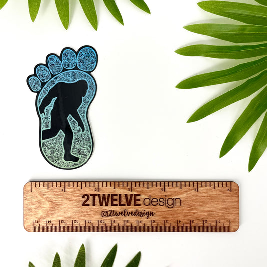 Intricate Bigfoot illustration made into a sticker. Dark mode color theme. Laying on top a white background with leaves and a rule to show actual size.