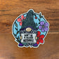 Sticker of a gnome holding a sign. Laying on a wood surface.