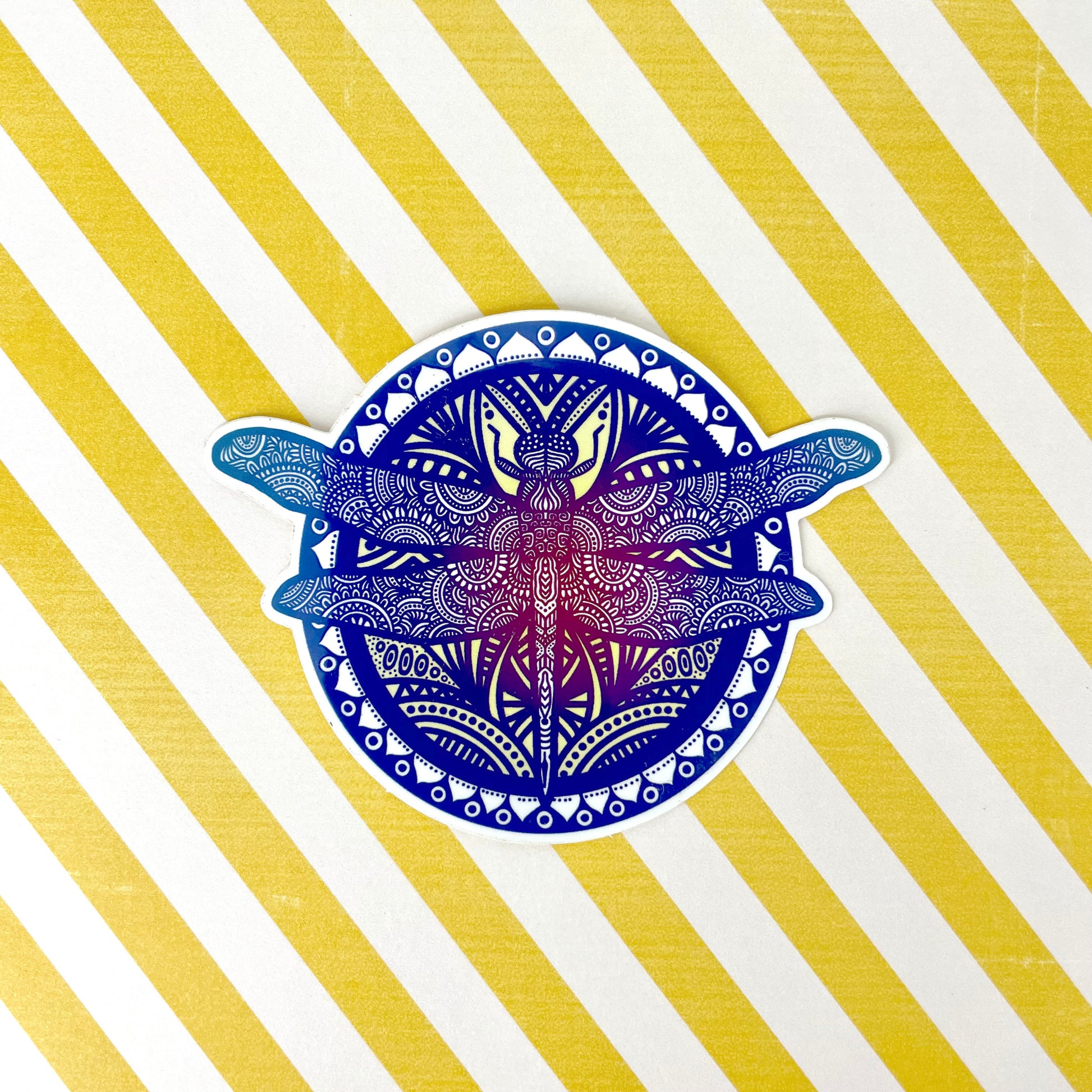 Beautiful mandala inspired dragonfly illustration made into a sticker. Laying on a white and yellow stripped background.