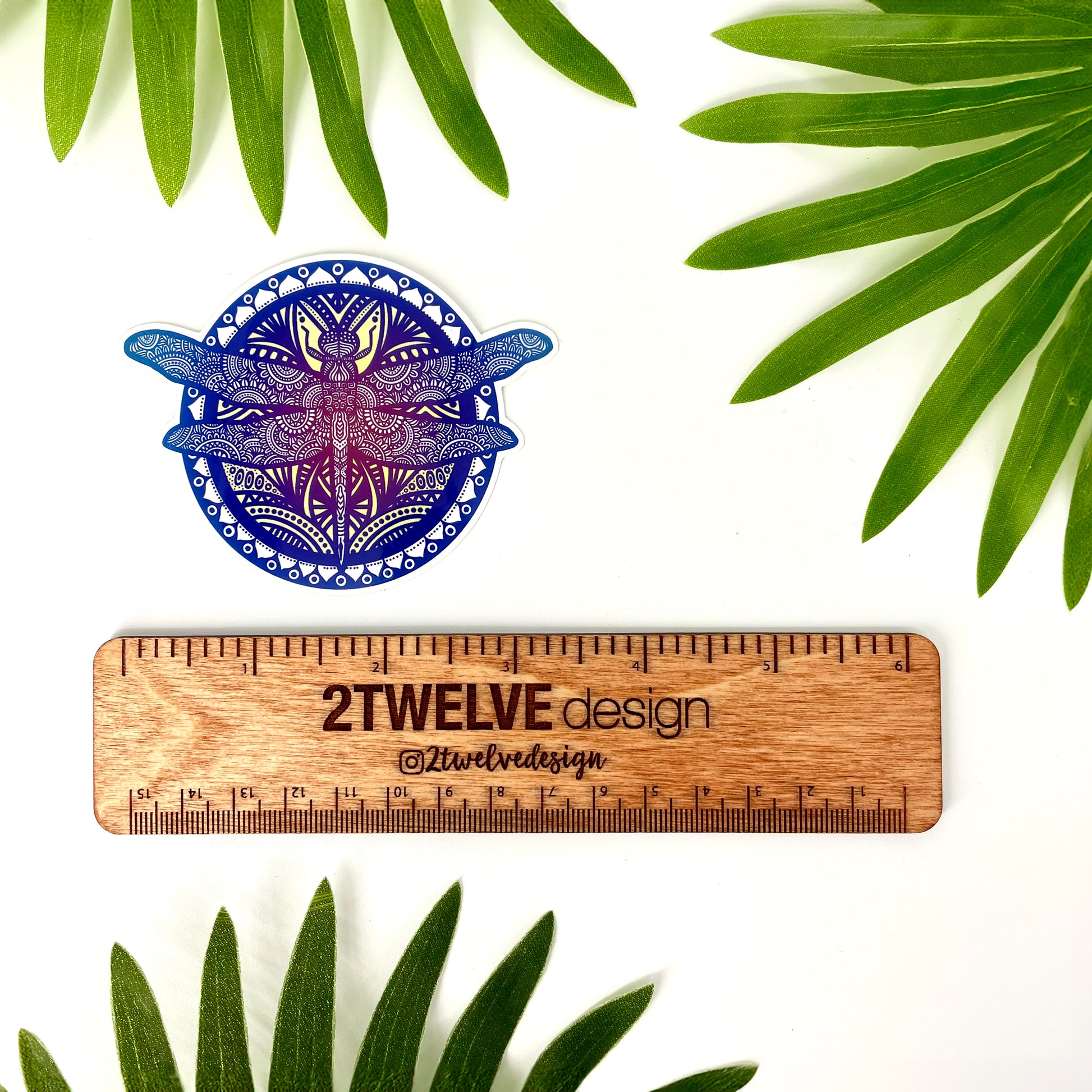 Wooden ruler with '2Twelve Design' branding and a blue and purple dragonfly illustrated sticker on a white background with green leaves.