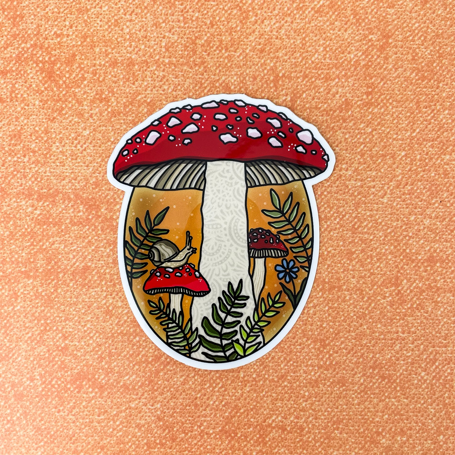 A die-cut sticker featuring an illustrated mushroom forest scene with red-capped mushrooms, green ferns, and a small snail, shown on a textured peach background.