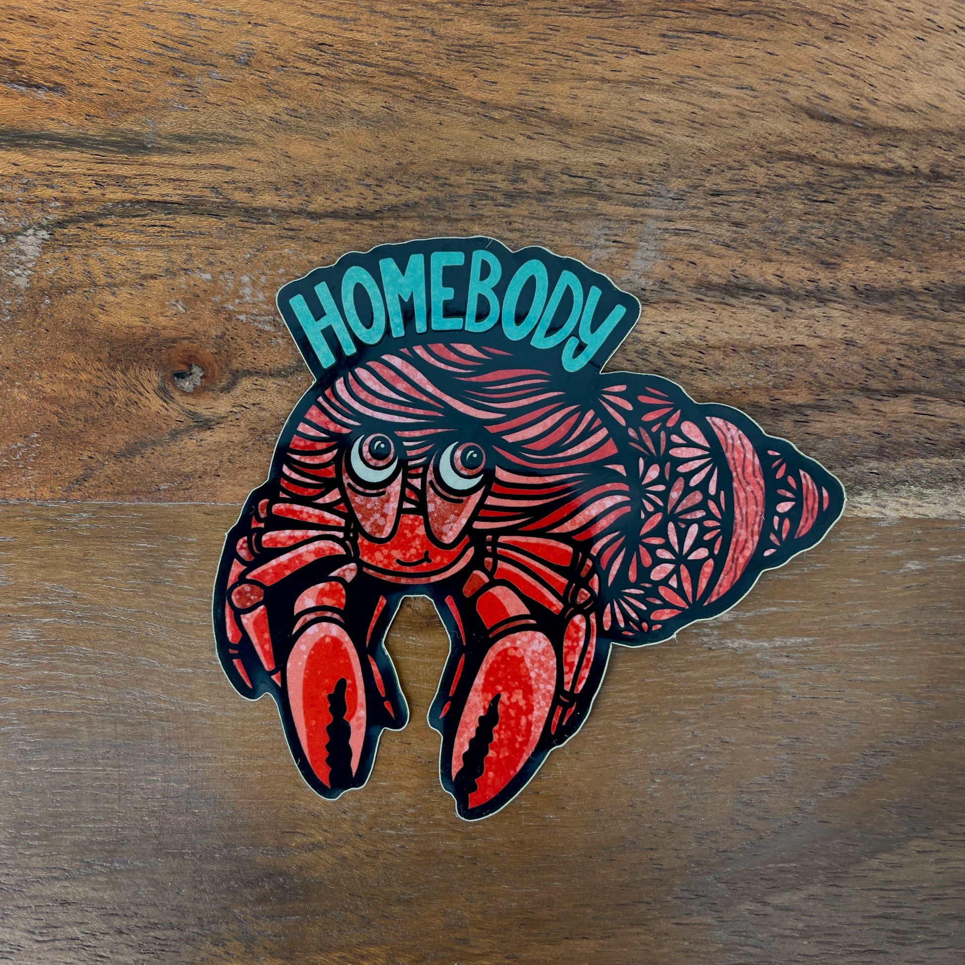 A hermit crab illustration made into a sticker with "Homebody" text. Laying on a wood surface.