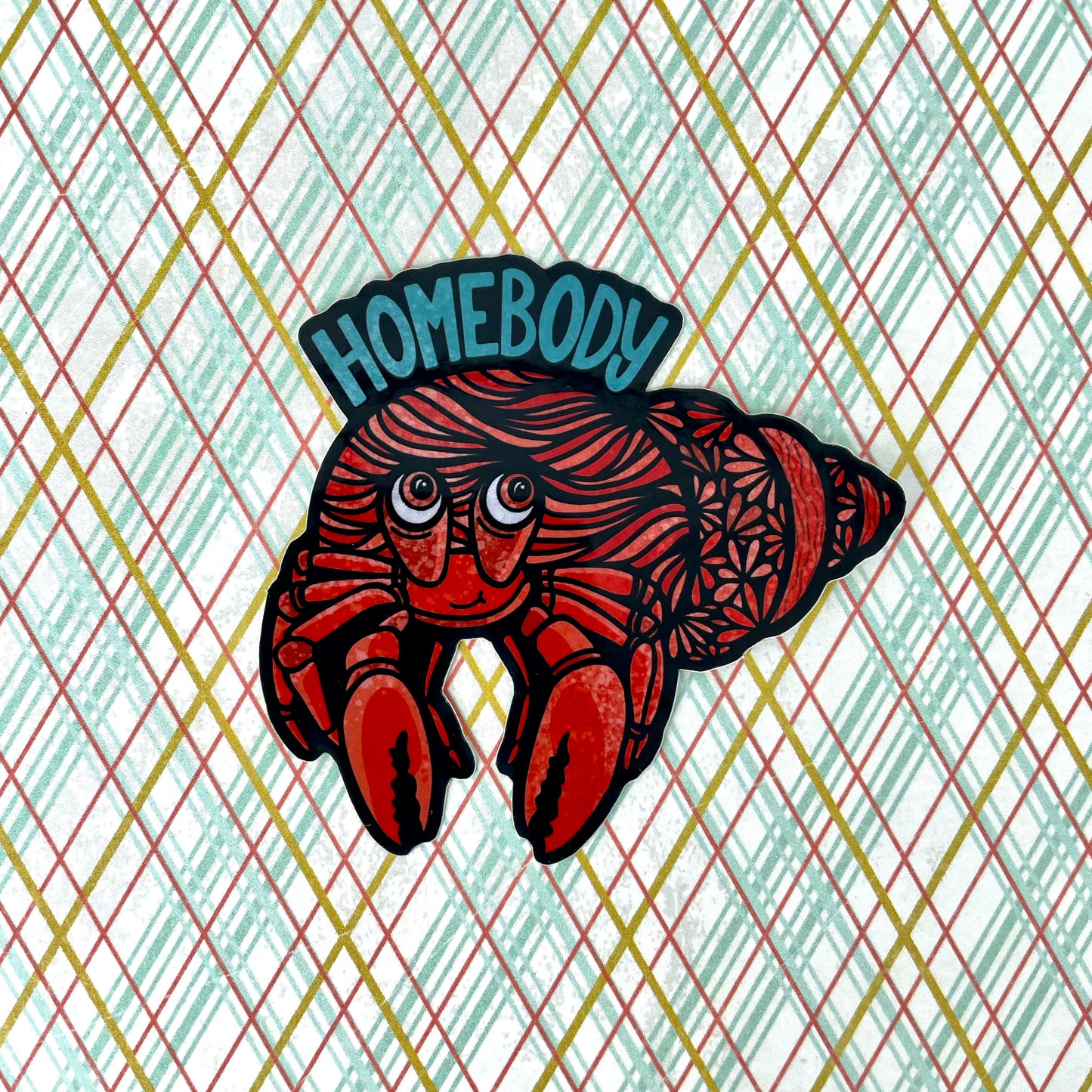 A sticker of a hermit crab with "Homebody" written above it. Laying on a colorful background.