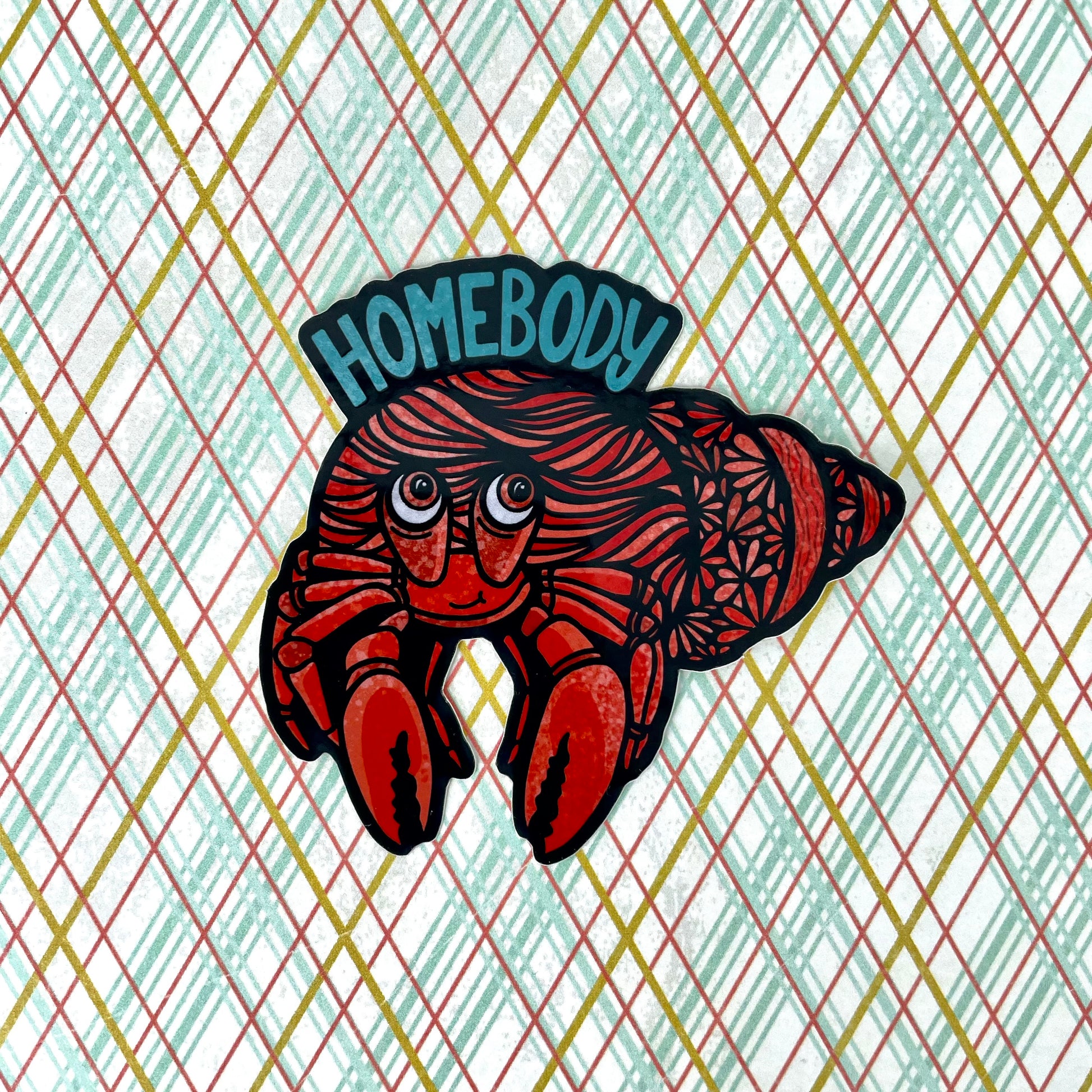 A sticker of a hermit crab with "Homebody" written above it. Laying on a colorful background.