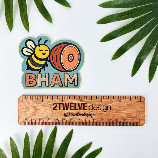 Sticker with bee and ham design labeled 'BHAM' next to a wooden ruler with '2Twelve Design' branding, surrounded by green leaves. Bellingham, Washington sticker.