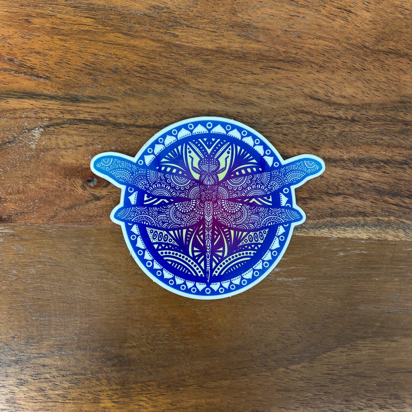 Blue and purple dragonfly mandala illustrated sticker laying on a wooden surface.
