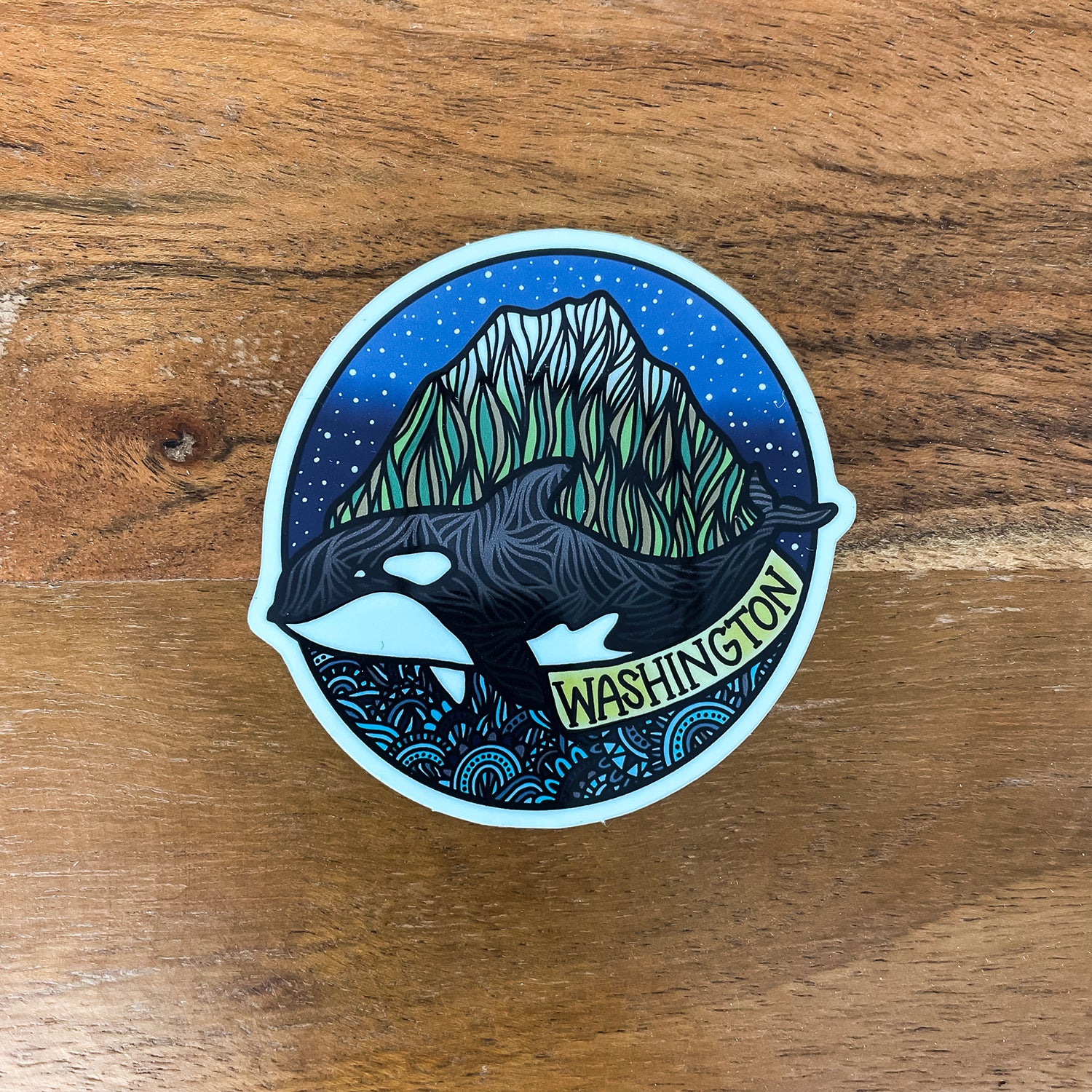 A circular sticker featuring an orca whale swimming in front of a mountain under a starry blue sky, with the word 'WASHINGTON' printed along the bottom edge, placed on a wooden surface.