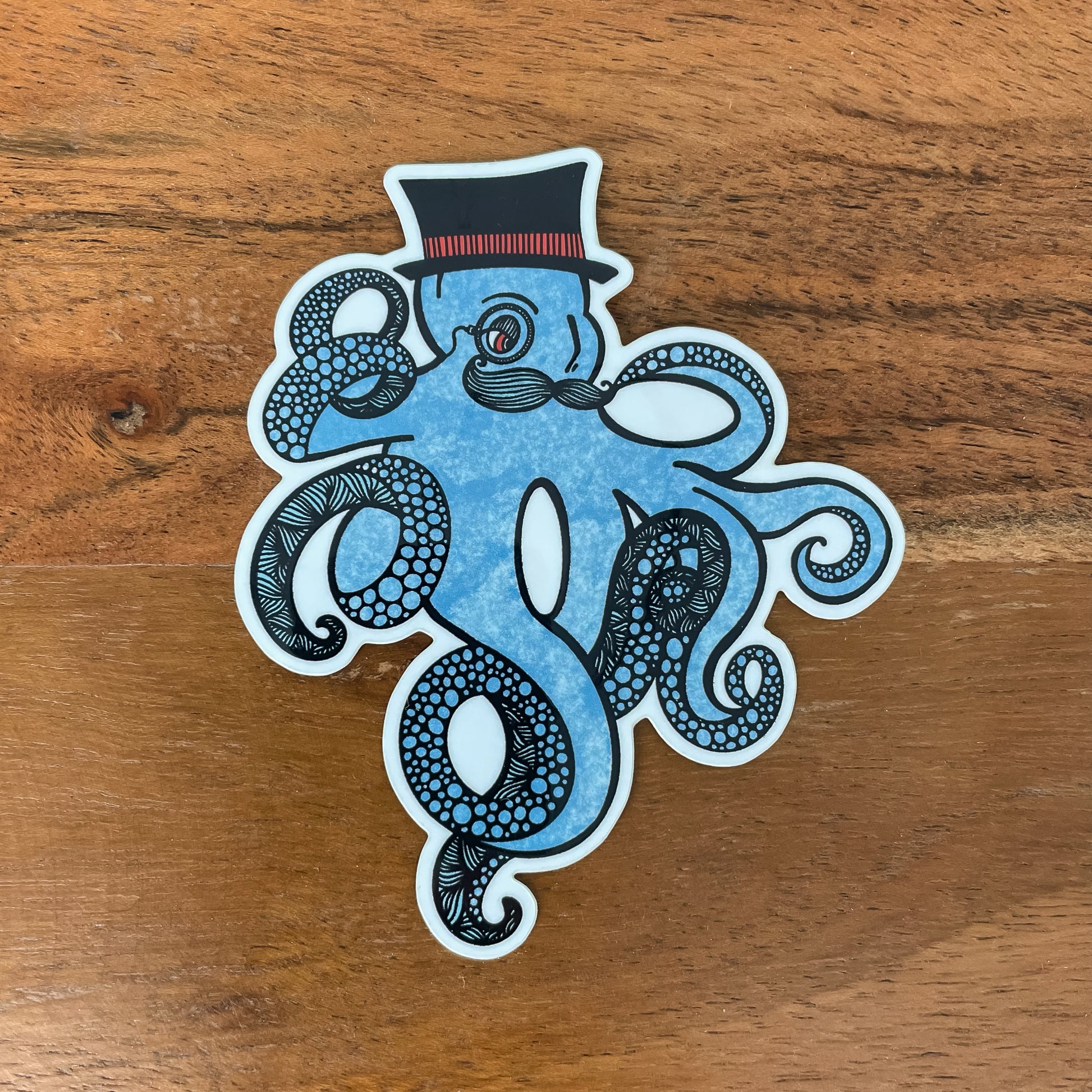 Blue kraken/octopus with a top hat and mustache. Detailed illustration made into a vinyl sticker laying on a wooden surface.